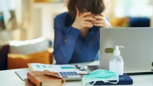 a woman exhibits stress with her head in her hands while at a desk with a laptop, hand sanitizer, and mask
