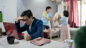an Asian man exhibits stress while working from home during a pandemic with children in the background