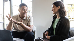 A workplace discussion between two people seated in chairs, one gesturing while the other listens and holds a tablet.