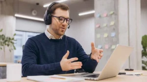 A person wearing a headset gestures with their hands while speaking at a desk with an open laptop in a modern office setting.