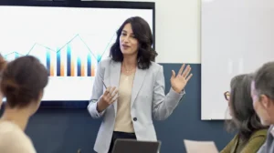 A woman presents data to colleagues during a business meeting, standing in front of a screen displaying a chart.