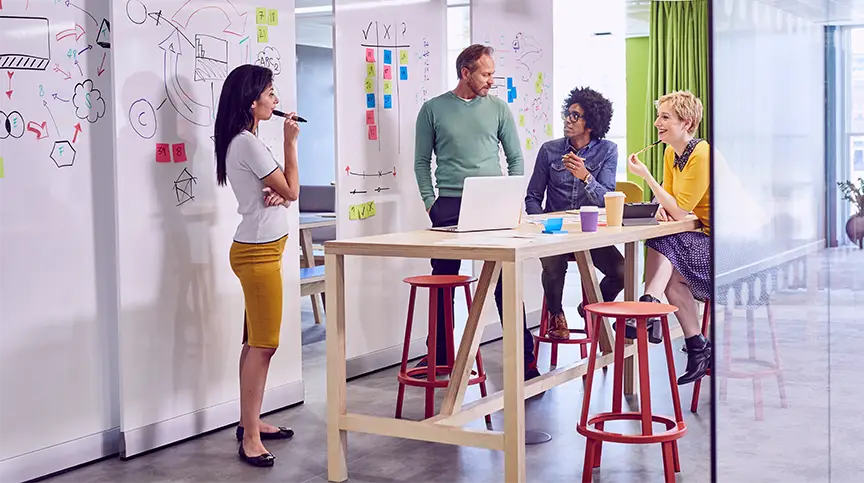 Group of people gathered around a high table with a laptop while brainstorming on whiteboards covered with notes and sketches.